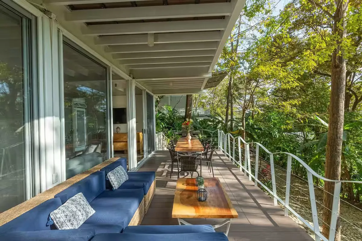 Long covered terrace with a blue sofa and dining table among the trees